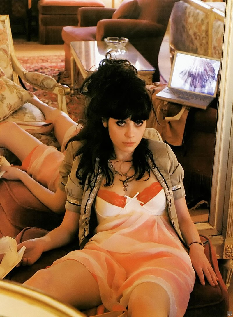 Zooey Deschanel lounges agains a mirror in orange dress with skirt hiked up to her thighs